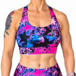 A close-up of a woman wearing a vibrant sports bra and matching high-waisted leggings set. The athletic wear features a bold, colorful abstract pattern with bright pink, electric blue, black, and white star and paint-splatter motifs.
