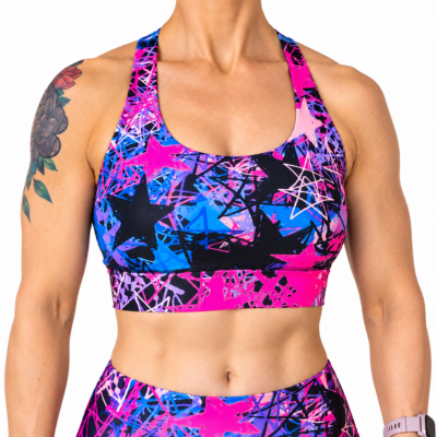 A close-up of a woman wearing a vibrant sports bra and matching high-waisted leggings set. The athletic wear features a bold, colorful abstract pattern with bright pink, electric blue, black, and white star and paint-splatter motifs.