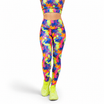 A person shown from the waist down wearing high-waisted leggings with a bright multicolour paint splash pattern in neon pink, yellow, green, blue, and purple, paired with neon yellow sneakers, against a plain white background.