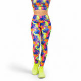 A person shown from the waist down wearing high-waisted leggings with a bright multicolour paint splash pattern in neon pink, yellow, green, blue, and purple, paired with neon yellow sneakers, against a plain white background.