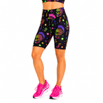 A model wearing high-waisted black athletic biker shorts with a vibrant neon multicolour sugar skull and paint splatter pattern, paired with bright pink running shoes against a plain white background.