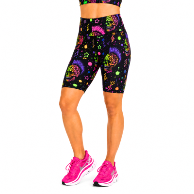 A model wearing high-waisted black athletic biker shorts with a vibrant neon multicolour sugar skull and paint splatter pattern, paired with bright pink running shoes against a plain white background.