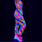 A model wearing a matching neon glow-print activewear set consisting of a sports bra and baggy pants, paired with glowing pink training shoes, shown against a solid black background under UV light.