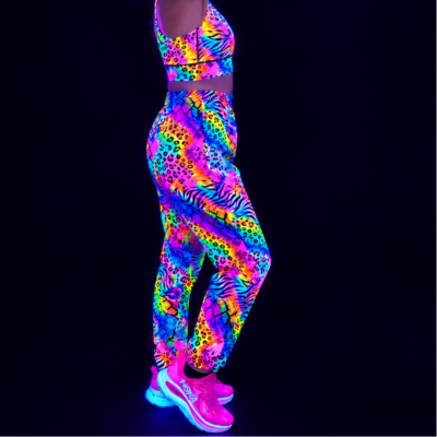 A model wearing a matching neon glow-print activewear set consisting of a sports bra and baggy pants, paired with glowing pink training shoes, shown against a solid black background under UV light.