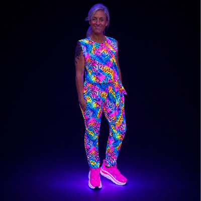 A woman stands against a black background wearing a sleeveless matching top and jogger pants set with a bright neon multicolour abstract print, paired with neon pink trainers that glow under UV light.