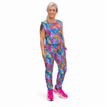 A woman modelling a sleeveless jumpsuit with a vibrant, multicoloured abstract pattern in shades of blue, pink, yellow, and purple, paired with bright pink trainers, against a plain white background.