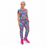 A woman modelling a sleeveless jumpsuit with a vibrant, multicoloured abstract pattern in shades of blue, pink, yellow, and purple, paired with bright pink trainers, against a plain white background.