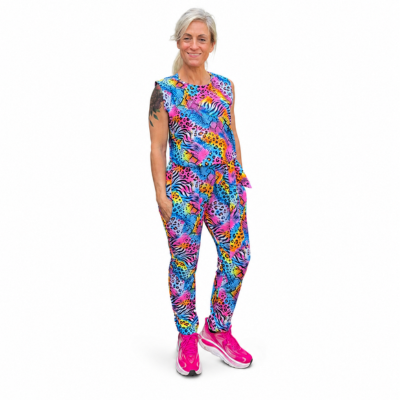 A woman modelling a sleeveless jumpsuit with a vibrant, multicoloured abstract pattern in shades of blue, pink, yellow, and purple, paired with bright pink trainers, against a plain white background.