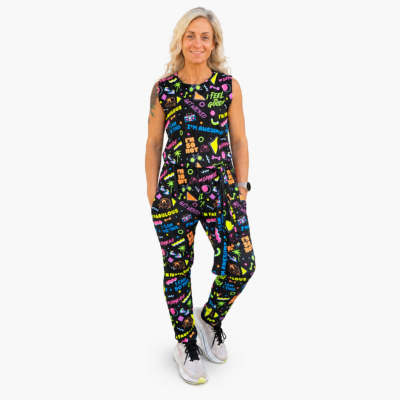 A woman modelling a sleeveless black jumpsuit with a colourful graffiti-style print, paired with white trainers, against a plain white background.
