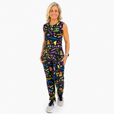 A woman modelling a sleeveless black jumpsuit with a vibrant multicolour neon-style graphic print, paired with white trainers, against a plain white background.