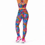A person shown from the back wearing a matching colourful athletic set with a bright multicolour abstract print, including a sports bra and high-waisted leggings, paired with pink trainers on a white studio background.