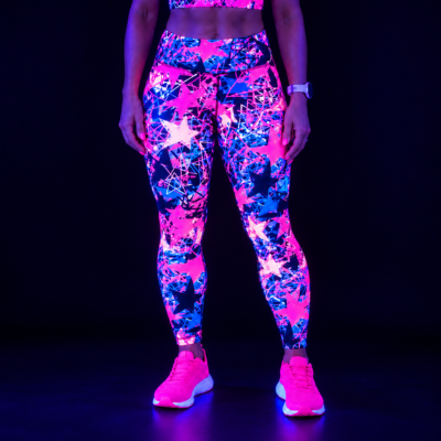 A model wearing high-waisted leggings and a matching sports bra with a glowing neon star and splatter pattern under blacklight, paired with bright pink running shoes, set against a dark background.