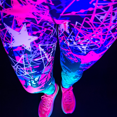 Close-up of a person's legs wearing neon UV-reactive leggings with a pink, blue, and white graffiti star pattern, paired with matching neon trainer, set against a solid black background.