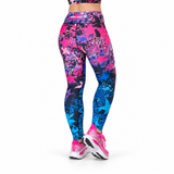 A person shown from the waist down wearing high-waisted fitness leggings with a bold abstract paint-splatter pattern in hot pink, electric blue, and black, paired with pink and white running shoes against a plain white background.