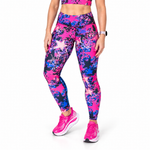 A model wearing high-waisted running tights with a vibrant pink, blue, and purple paint-splatter pattern, paired with matching pink training shoes, shown against a plain white background.