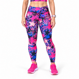 A model wearing high-waisted fitness leggings with a vibrant pink, blue, and black star and splatter pattern, paired with matching pink trainers, shown against a plain white background.