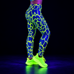 A person wearing high-waisted leggings with a fluorescent neon green giraffe-like pattern on a bright blue base, paired with glowing neon yellow running shoes, photographed against a black background.