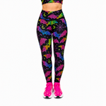 A person wearing high-waisted leggings with a black background and a colourful neon pattern of bats, moons, and stars, paired with bright pink trainers against a plain white background.