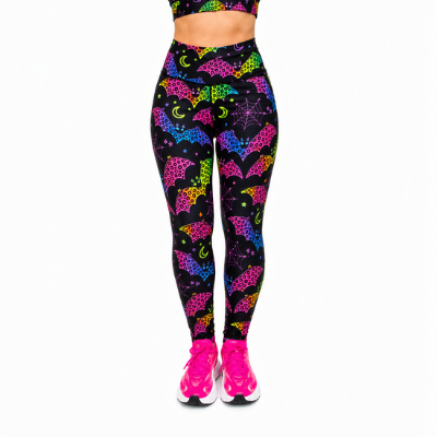 A person wearing high-waisted leggings with a black background and a colourful neon pattern of bats, moons, and stars, paired with bright pink trainers against a plain white background.