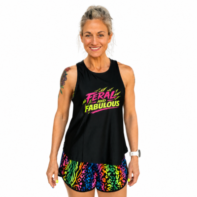A woman modeling a black sleeveless tank top with a colorful graphic that reads 'FERAL AND FABULOUS' in neon pink, yellow, and green, paired with neon patterned shorts, against a plain white background.