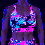 A close-up of a person wearing a neon UV-reactive sports bra and matching bottoms with a bright pink, blue, and black star pattern, photographed under blacklight against a dark background.
