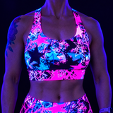 A close-up of a person wearing a neon UV-reactive sports bra and matching bottoms with a bright pink, blue, and black star pattern, photographed under blacklight against a dark background.