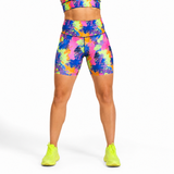 A model from the waist down wearing high-waisted athletic shorts and a matching sports bra with a vibrant multicolour paint plash pattern, paired with neon yellow trainers, against a plain white background.
