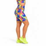 A model wearing high-waisted fitness shorts and a matching sports bra with a vibrant multicolour paint splash pattern, paired with neon yellow trainers, shown from a side-back angle on a white background.