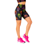 A model wearing high-waisted black biker shorts with a colourful sugar skull pattern, paired with bright pink running shoes, shown from the waist down on a white background.