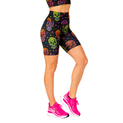 A model wearing high-waisted black biker shorts with a colourful sugar skull pattern, paired with bright pink running shoes, shown from the waist down on a white background.
