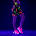 A model from the waist down wearing black fitness shorts with a vibrant, multicoloured neon sugar skull pattern under UV light, paired with bright pink trainers.