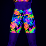A person wearing neon tie-dye athletic shorts and a matching crop top under blacklight, showing bright fluorescent purple, blue, green, yellow, orange, and pink patterns against a black background.