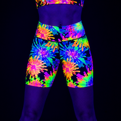 A person wearing neon tie-dye athletic shorts and a matching crop top under blacklight, showing bright fluorescent purple, blue, green, yellow, orange, and pink patterns against a black background.