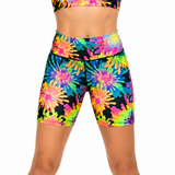A model wearing high-waisted athletic shorts with a vibrant neon tie-dye burst pattern on a black base, paired with a matching sports bra against a plain white background.