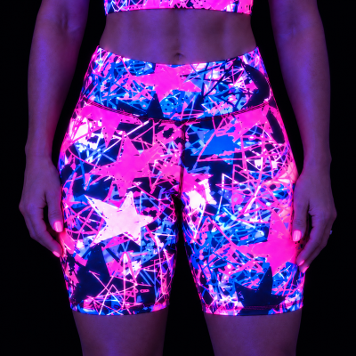 A person wearing high-waisted fitness shorts with a fluorescent neon star and geometric pattern in glowing pink, blue, and purple, photographed against a black background under UV lighting.