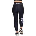 Rear view of a person wearing black high-waisted athletic leggings with a white 'WILD LEGS' logo on the waistband and a white graphic print on the lower right leg, paired with black trainers against a white background.