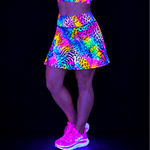 A model wearing a vibrant, multicolour UV-reactive athletic skirt with neon patterns, paired with neon pink trainers, shown against a solid black background under blacklight.