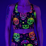 A person wearing a black UV-reactive sports bra and matching high-waisted shorts set with a vibrant neon sugar skull and star pattern, shown under blacklight against a purple background.