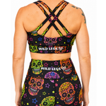 Rear view of a model wearing a matching black activewear set with a vibrant multicolor sugar skull and star pattern. The set includes a sports bra with crisscross straps and a high-waisted athletic skirt, both featuring the 'WILD LEGS' brand name.
