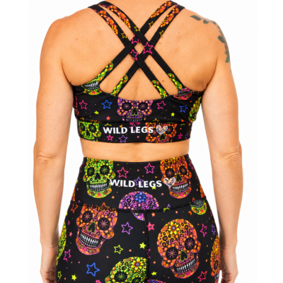 Rear view of a model wearing a matching black activewear set with a vibrant multicolor sugar skull and star pattern. The set includes a sports bra with crisscross straps and a high-waisted athletic skirt, both featuring the 'WILD LEGS' brand name.