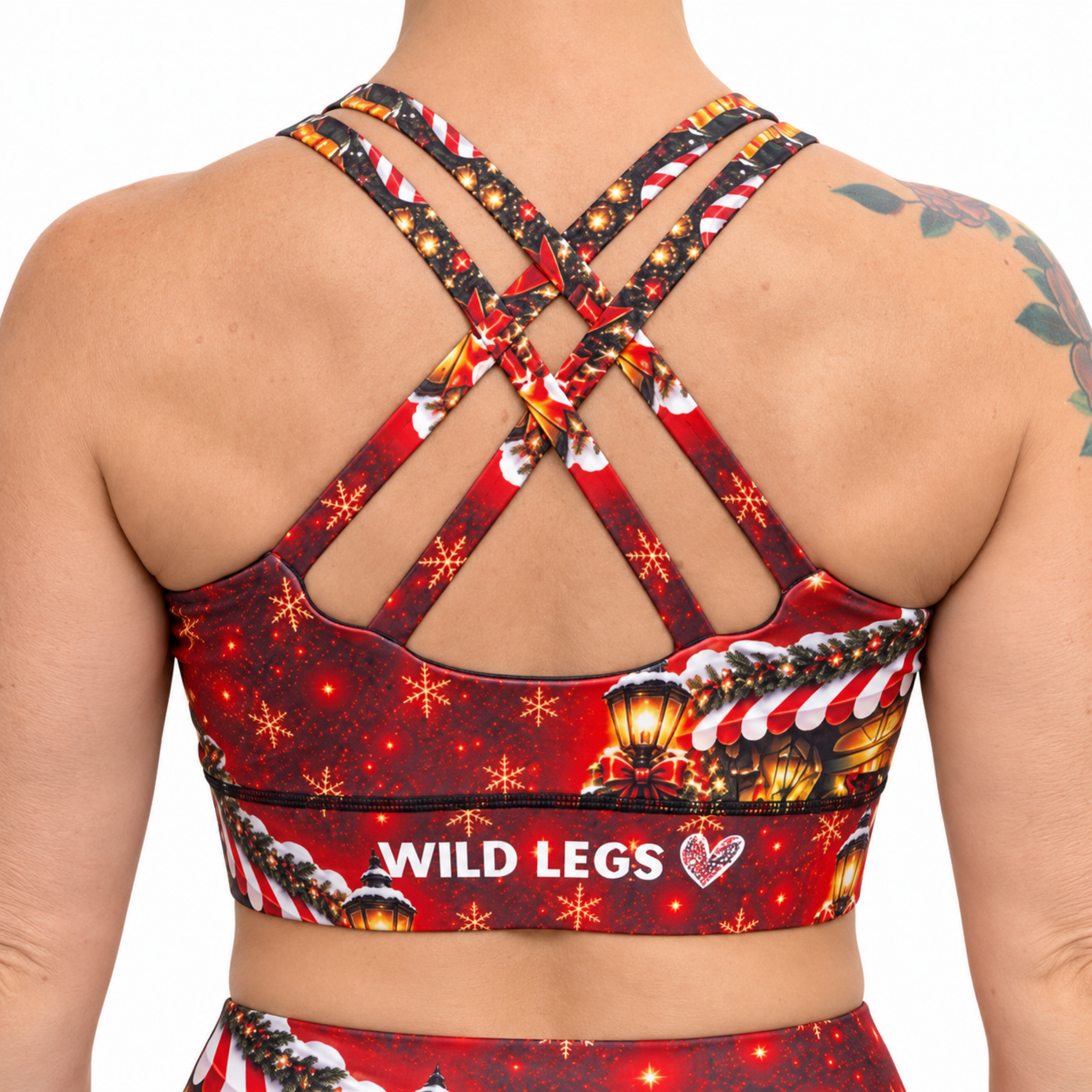 The back view of a sports bra in a Christmas design in red, with gold snow covered lanterns, a Christmas house and the Wild Legs logo on the band, on a white background