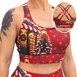 A woman wearing a red Christmas-themed sports bra and matching bottoms, with a circular inset showing the back of the sports bra. The sports bra features a festive print with a lit holiday house, Christmas tree, candy canes, and snowflakes on a red background.