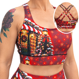 A woman wearing a red Christmas-themed sports bra and matching bottoms, with a circular inset showing the back of the sports bra. The sports bra features a festive print with a lit holiday house, Christmas tree, candy canes, and snowflakes on a red background.