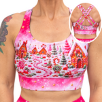 A woman wearing a pink sports bra with a colorful gingerbread village print, shown from the chest down with a circular inset highlighting the back straps and band text that reads 'WILD LEGS'.