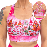 A woman wearing a pink sports bra with a colorful gingerbread village print, shown from the chest down with a circular inset highlighting the back straps and band text that reads 'WILD LEGS'.