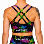 Rear view of a person wearing a black sports bra and matching bottoms with a bright neon multicolor print of bats, moons, and stars. The sports bra has crisscross back straps and the brand name 'WILD LEGS' is printed in white on the back.