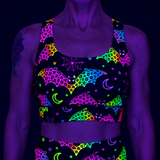 A black UV-reactive sports bra and matching bottoms set with a neon multicolor print of bats, crescent moons, stars, and spiderwebs, shown on a model under blacklight.