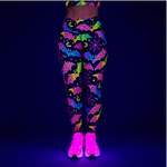 A person wearing high-waisted leggings and glowing neon trainers under blacklight, shown from the waist down against a dark blue background. The leggings have a black base with bright neon multicolor patterns including bats, moons, and stars.