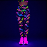 A person wearing high-waisted leggings and glowing neon trainers under blacklight, shown from the waist down against a dark blue background. The leggings have a black base with bright neon multicolor patterns including bats, moons, and stars.