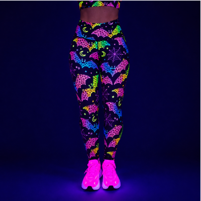 A person wearing high-waisted leggings and glowing neon trainers under blacklight, shown from the waist down against a dark blue background. The leggings have a black base with bright neon multicolor patterns including bats, moons, and stars.